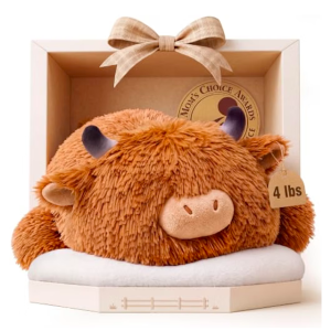 A plush, light brown, shaggy-furred Highland cow toy resting on a white, cloud-like cushion inside an open, light-beige hexagonal display box. The cow has short, smooth gray horns, a flat plush snout, and small dark eyes. A small brown tag hangs from its right horn reading "4 lbs". A simple checkered bow sits atop the display case, which features a golden circle logo reading "Mom's Choice Awards."