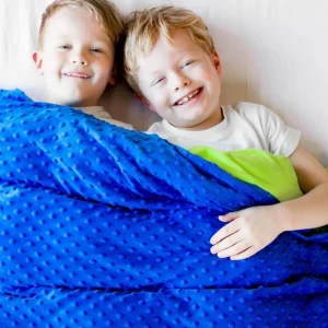 Two young boys lying side-by-side on a bed, smiling happily while tucked under a vibrant blue weighted blanket. The blanket features a textured, bumpy minky dot pattern on the exterior and rolls down slightly to reveal a bright lime-green underside near the boy on the right. Both children are wearing white t-shirts against light-colored bedding.