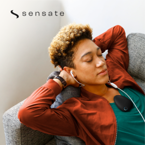 A person with short, curly blonde and brown hair reclining comfortably with closed eyes on a gray textured sofa, wearing white wired earbuds and a rust-orange jacket over a teal shirt. Resting on the center of their chest is the Sensate, a smooth, matte-black, pebble-shaped relaxation device with a single glowing blue circular light indicator. The upper-left corner of the image displays the brand logo—a stylized black "S" next to the word "sensate" in lowercase text.